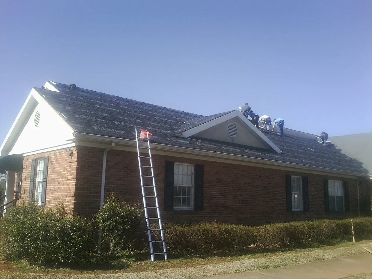 Crew tearing shingles off a brick ranch roof with a ladder set — Emergency Roof Repair work by our crew in Danciger